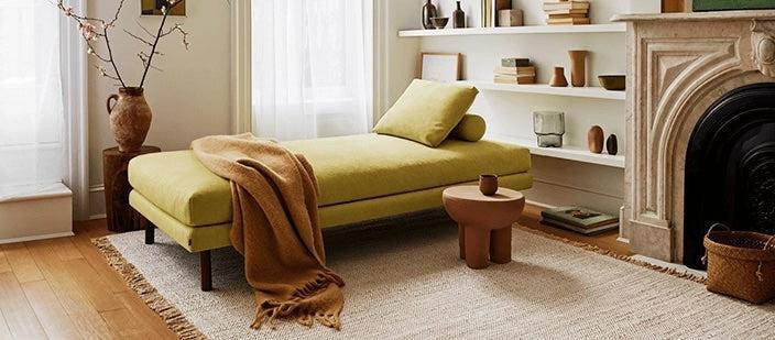 A cozy living room features a yellow daybed with a brown throw blanket, a small rustic wooden table, a white bookshelf with various decorative items, and a fireplace with a stone mantel. Sunlight filters through sheer white curtains, illuminating the wooden floor.
