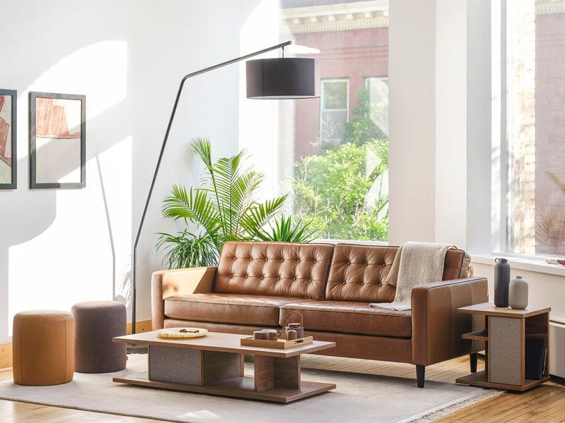 A modern living room with a brown leather tufted sofa, a wooden coffee table, and a standing floor lamp with a black shade. There are two poufs, framed art on the walls, and a large window letting in natural light, revealing greenery outside.