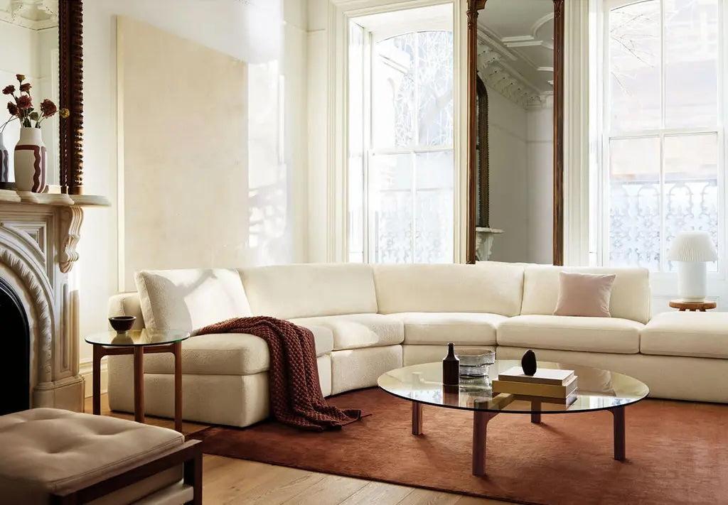 A cozy living room featuring a large, white sectional sofa adorned with a red throw blanket and a pink cushion. Two round glass coffee tables hold books, vases, and a drink. Tall windows allow natural light to flood the space, highlighting the ornate fireplace and mirror.
