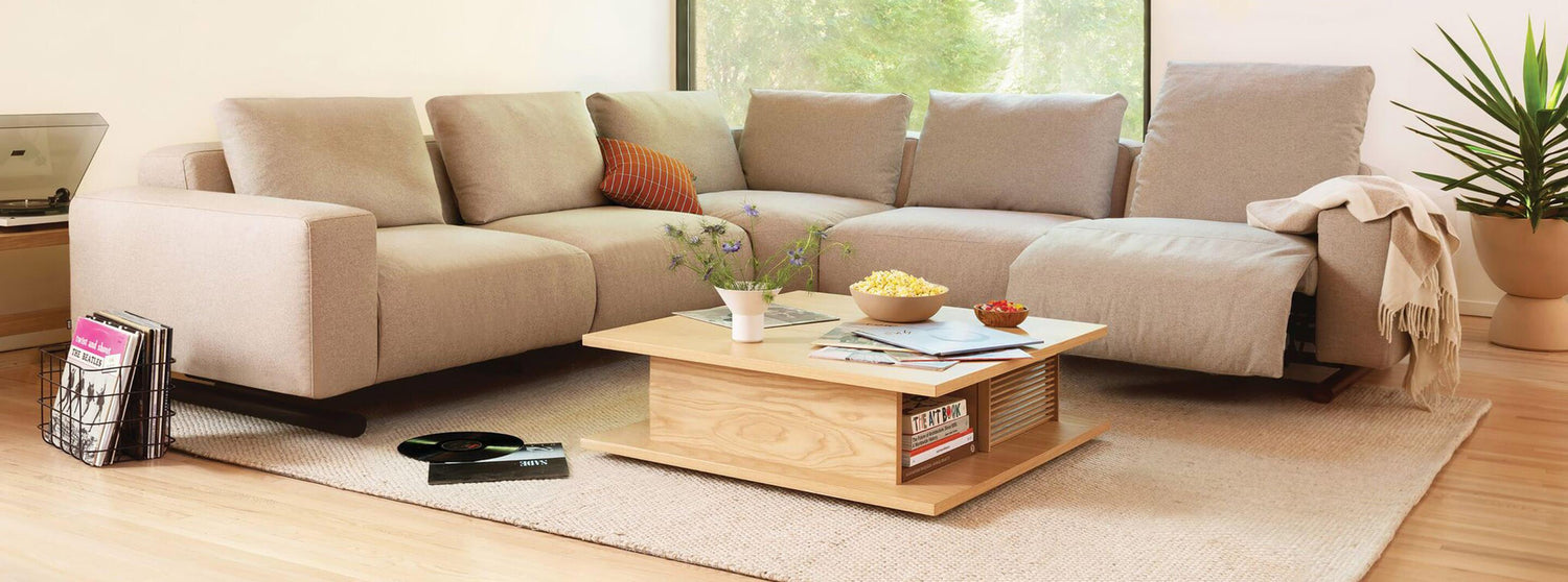 A cozy living room features a light beige sectional sofa adorned with assorted throw pillows and a beige blanket. A wooden coffee table holds a bowl of popcorn, a book, and a plant. Greenery is seen outside the large window, and a potted plant sits in the right corner.