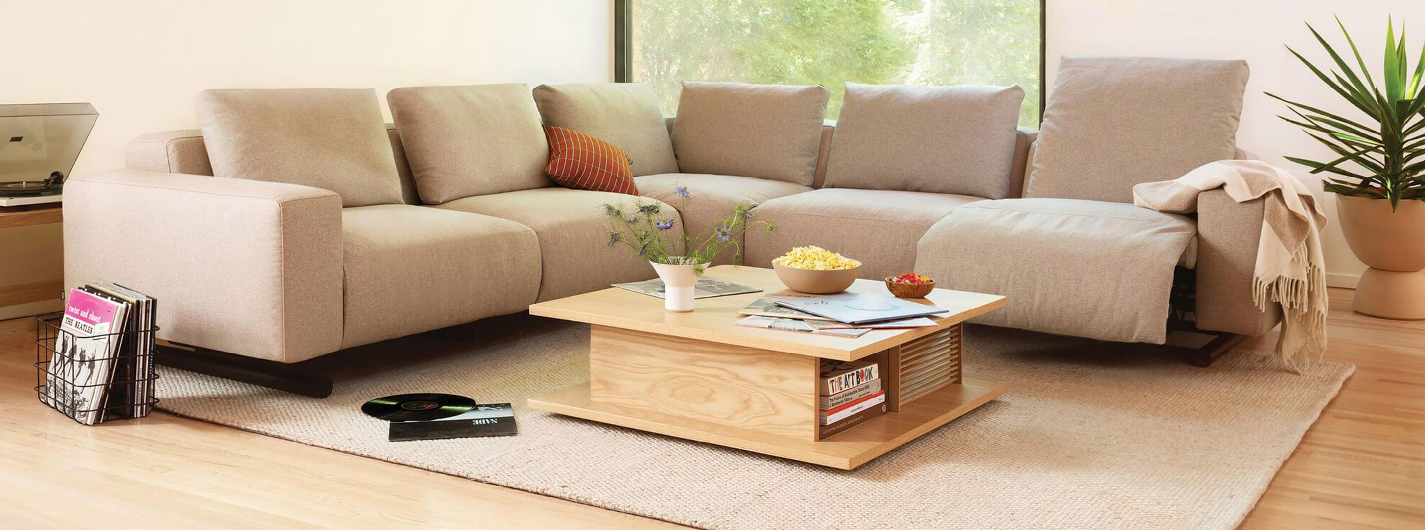 A cozy living room features a light beige sectional sofa adorned with assorted throw pillows and a beige blanket. A wooden coffee table holds a bowl of popcorn, a book, and a plant. Greenery is seen outside the large window, and a potted plant sits in the right corner.