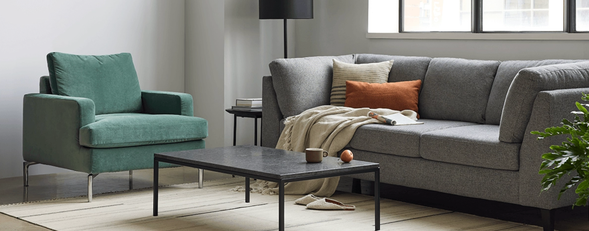 A modern living room featuring a gray sofa with an orange and beige cushion, and a light throw blanket. A green armchair sits nearby with a black floor lamp behind it. A rectangular black coffee table holds decorative items, and a large window lets in light.