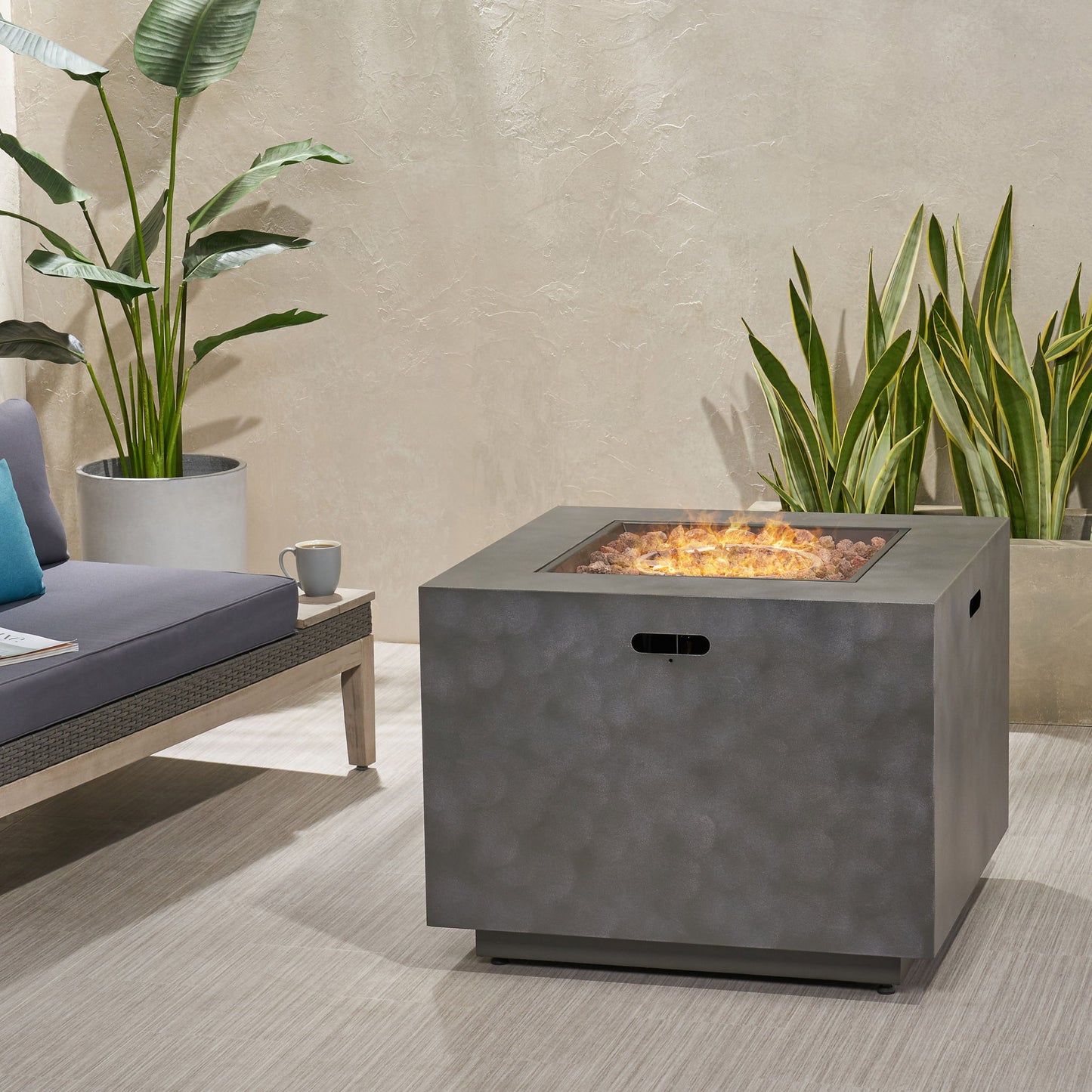 A modern patio features a gray sofa, two potted plants, and the Outdoor Patio Gas Burning Fire Pit Table 50,000 Btu, Square with visible flames. The scene is set against a light beige textured wall.