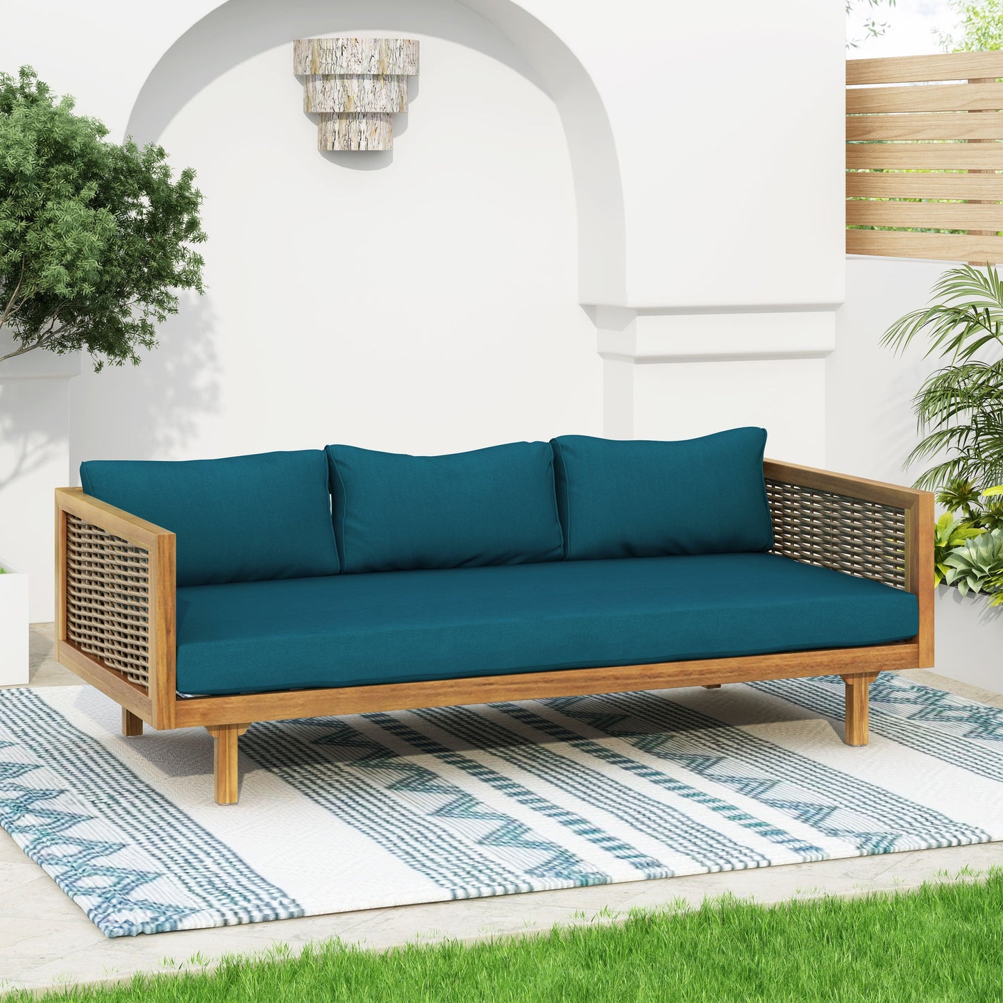 The Claremont 3 Seater Daybed in teak acacia wood with water resistant teal cushions sits on a blue and white patterned rug, surrounded by green plants, a white wall with arched niche, and a wooden privacy screen.