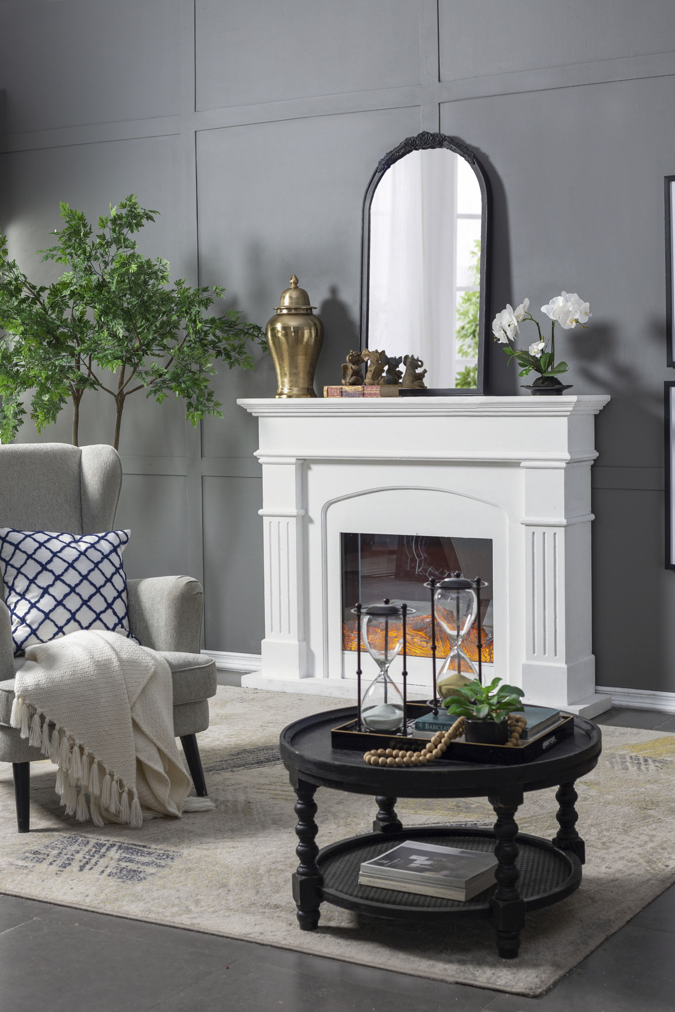 A modern living room features a white fireplace topped with the Hand Carved Rose Antique Mirror Frame, potted plants, a gold vase, gray armchair with blanket, and a round black coffee table on a beige rug.