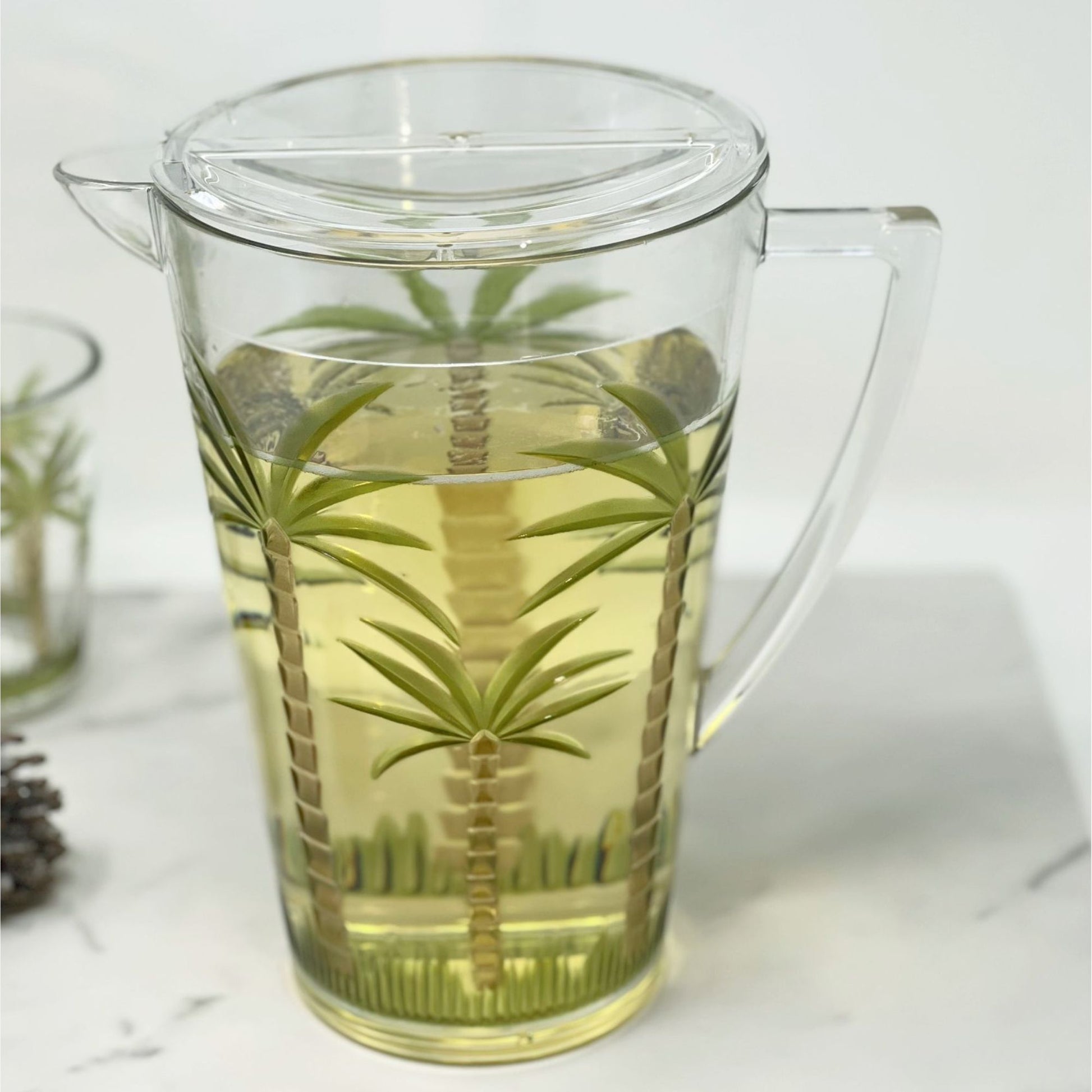 The Unbreakable Plastic Water Pitcher With Lid, Palm Tree Design - Clear, filled with a light yellow drink, sits on a white surface. A matching glass and a pinecone are partially visible nearby.