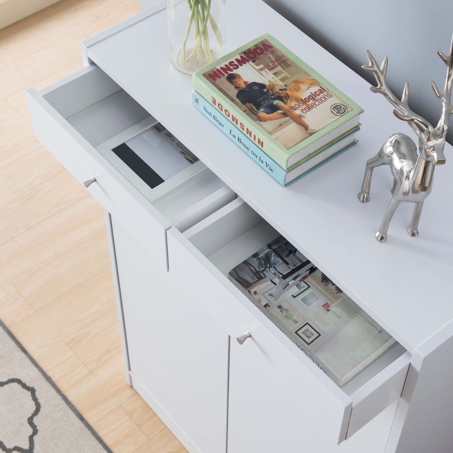A white shoe storage cabinet with five shelves and two open drawers for magazines, topped with stacked books and a silver deer figurine on a light wood floor; holds up to 15 pairs of shoes.