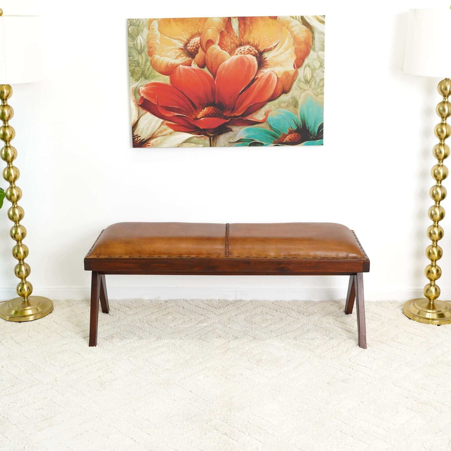 The Chad Mid-Century Modern Leather Bench in brown/tan rests on a light carpet between two gold floor lamps with round bases, beneath a colorful floral painting on a white wall.