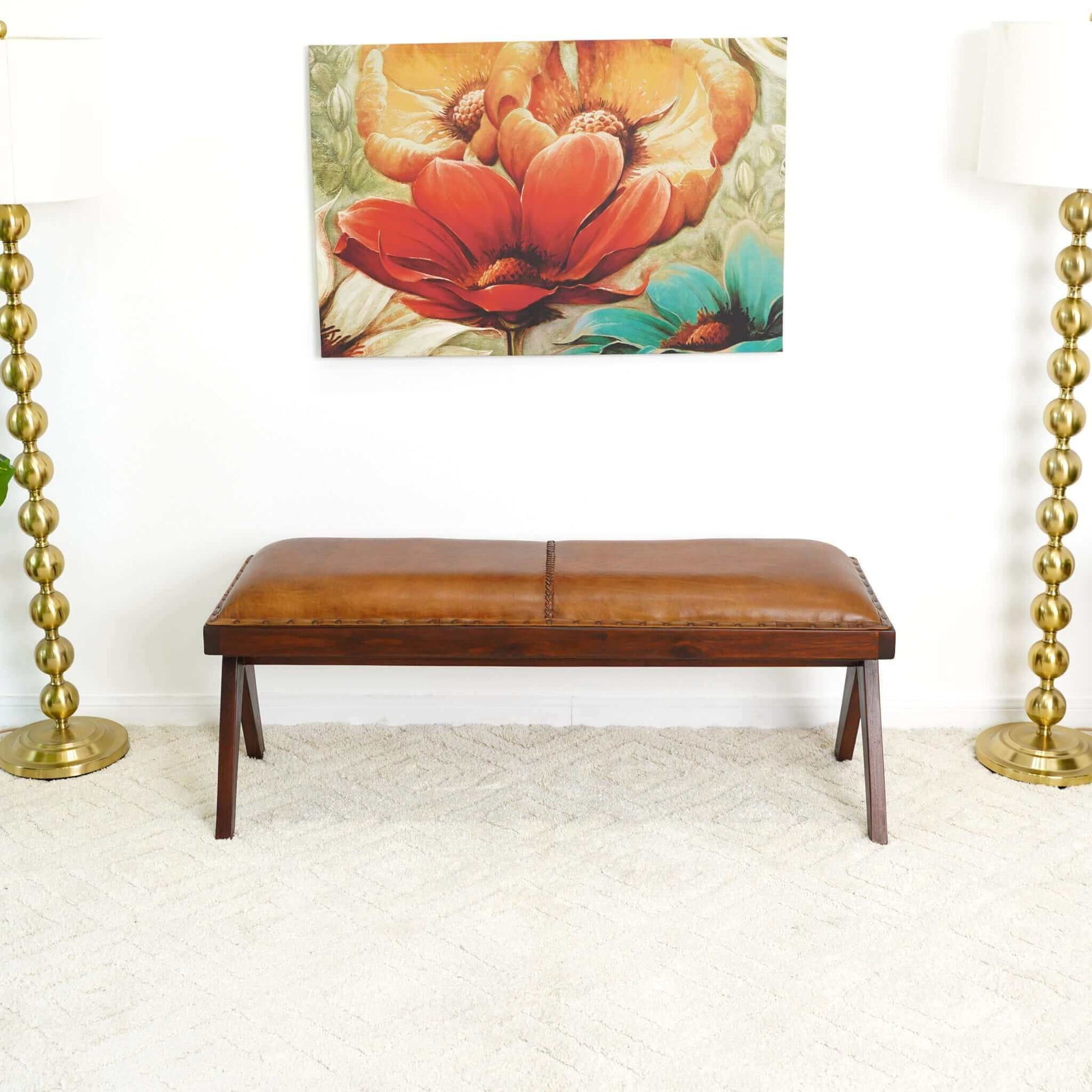The Chad Mid-Century Modern Leather Bench in brown/tan rests on a light carpet between two gold floor lamps with round bases, beneath a colorful floral painting on a white wall.