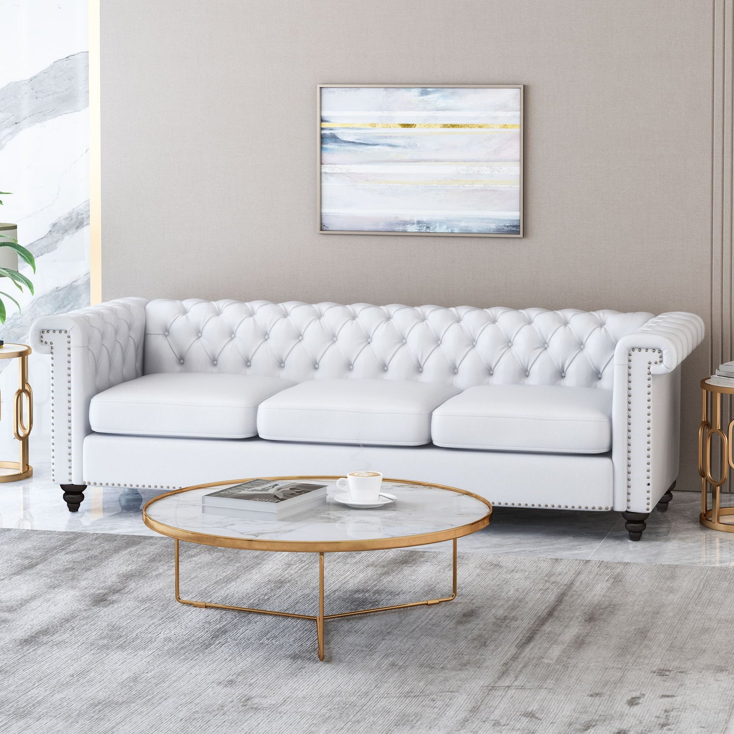 A modern living room with a Chesterfield Style 3 Seater Sofa with deep button tufting, a round marble coffee table with gold legs, gray rug, and framed abstract art on a beige wall. A cup and magazine are on the coffee table.