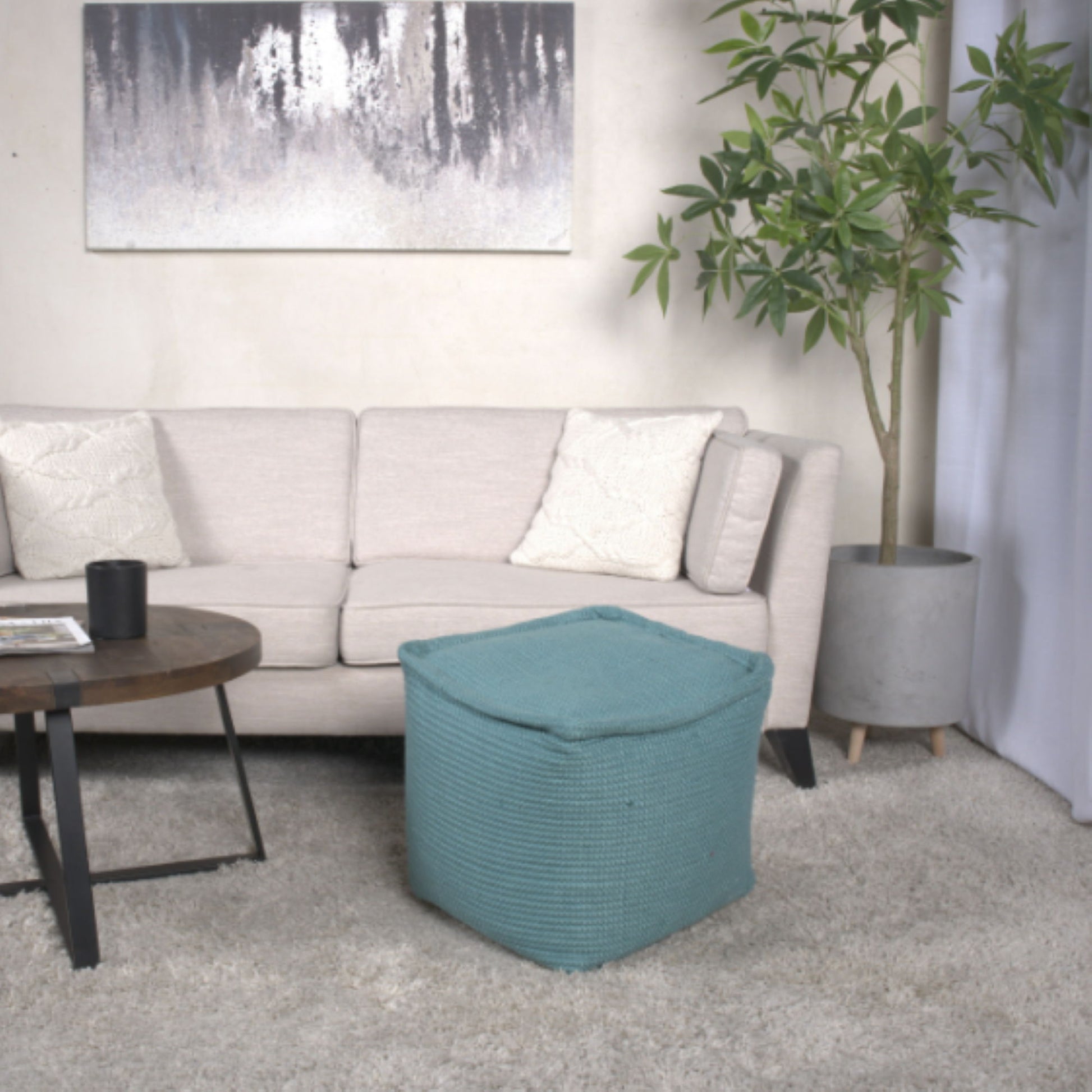 A modern living room featuring a light beige sofa with two white pillows, a Hansolo Teal Yarn Square Pouf Ottoman as a floor cushion, a round wooden coffee table, a large potted plant, and abstract wall art above the sofa.
