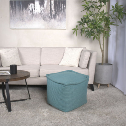 A modern living room featuring a light beige sofa with two white pillows, a Hansolo Teal Yarn Square Pouf Ottoman as a floor cushion, a round wooden coffee table, a large potted plant, and abstract wall art above the sofa.