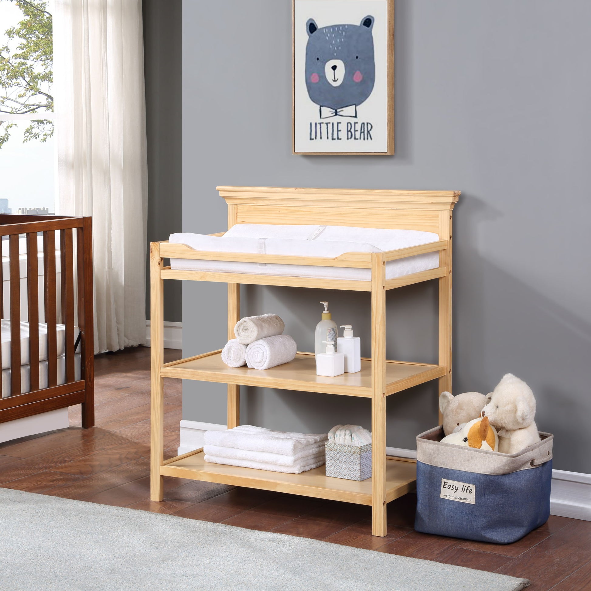 The Universal - Changing Table, topped with towels, baby supplies, and stuffed animals, stands in a nursery. Above it is a framed "Little Bear" picture, with a crib and window shown in the background.