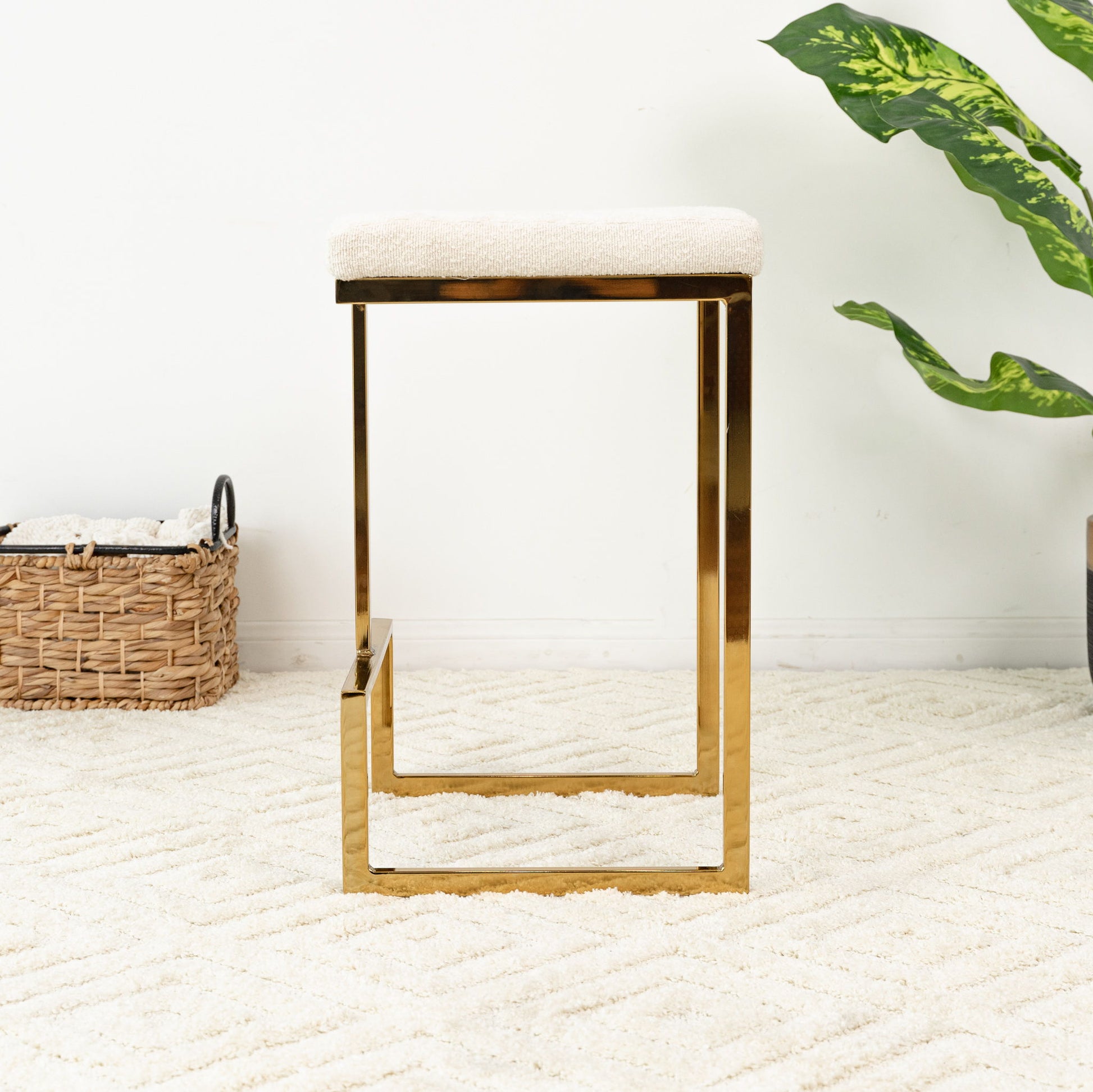 The Joel - Mid Century Modern Luxury Upholstered Stool, featuring a gold metal frame and white cushioned seat, rests on a textured cream rug. In the background, towels in a woven basket and a leafy green plant sit against a white wall.
