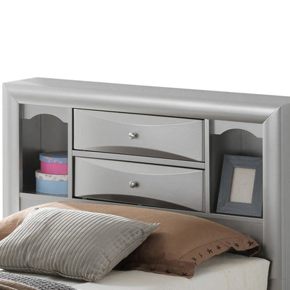 The Marilla Storage Bed with Bookcase Headboard features a gray headboard with two central drawers, open side shelves for storage or display, placed behind a bed dressed in brown and striped pillows and a beige sheet.