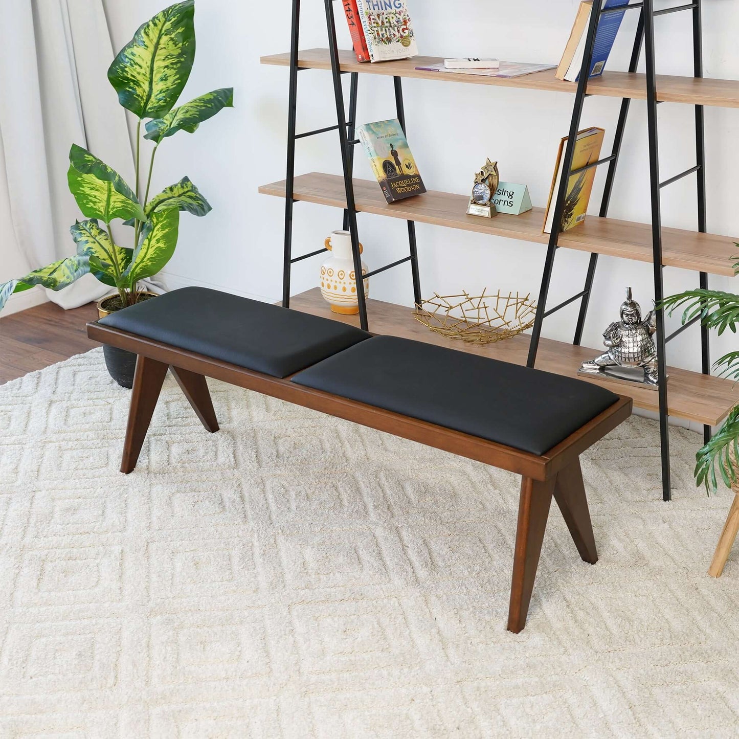 The Keira - Modern Aesthetics Bench with black cushions sits on a light textured rug, surrounded by green potted plants and a shelving unit displaying books, décor, and a Buddha statue.