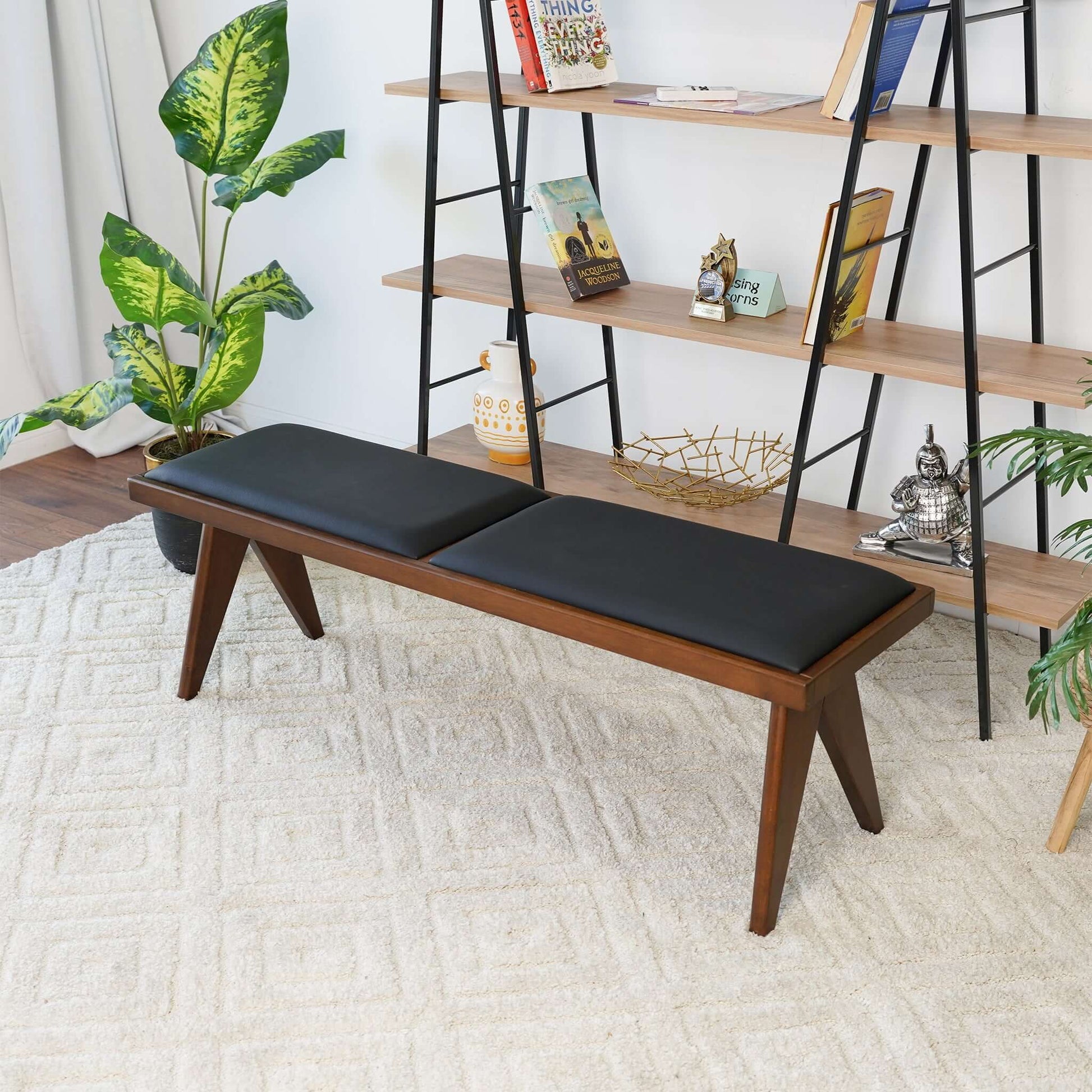 The Keira - Modern Aesthetics Bench with black cushions sits on a light textured rug, surrounded by green potted plants and a shelving unit displaying books, décor, and a Buddha statue.