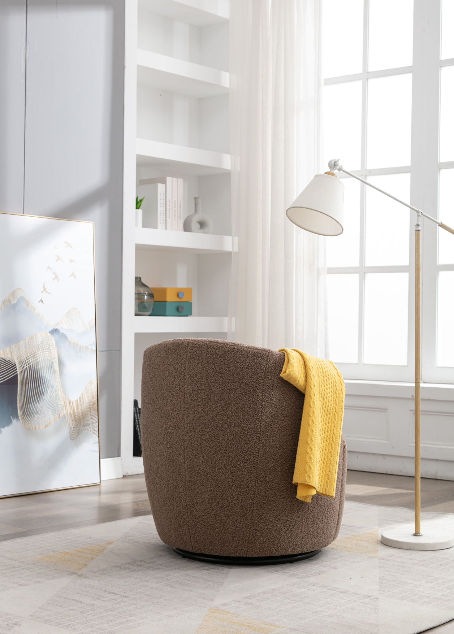 The Teddy Fabric Swivel Accent Armchair Barrel Chair, in cozy brown with a yellow blanket, sits by a tall window near a white floor lamp, built-in shelves, and abstract artwork in a bright room.