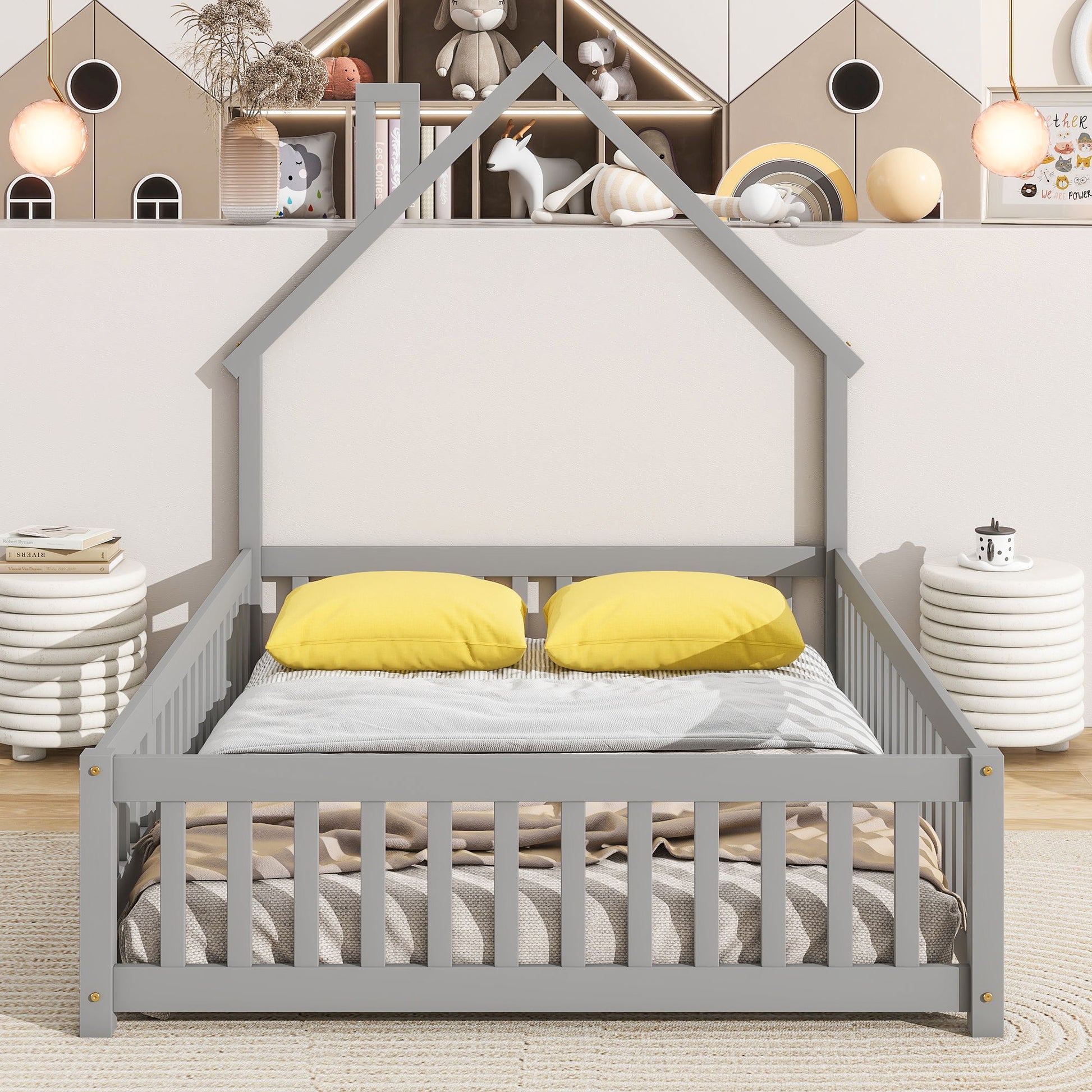 The House-Shaped Headboard Floor Bed With Fence features a gray frame, two yellow pillows, and a beige blanket in a cozy room with soft lighting, round side tables, and wall shelves decorated with toys and books behind it.