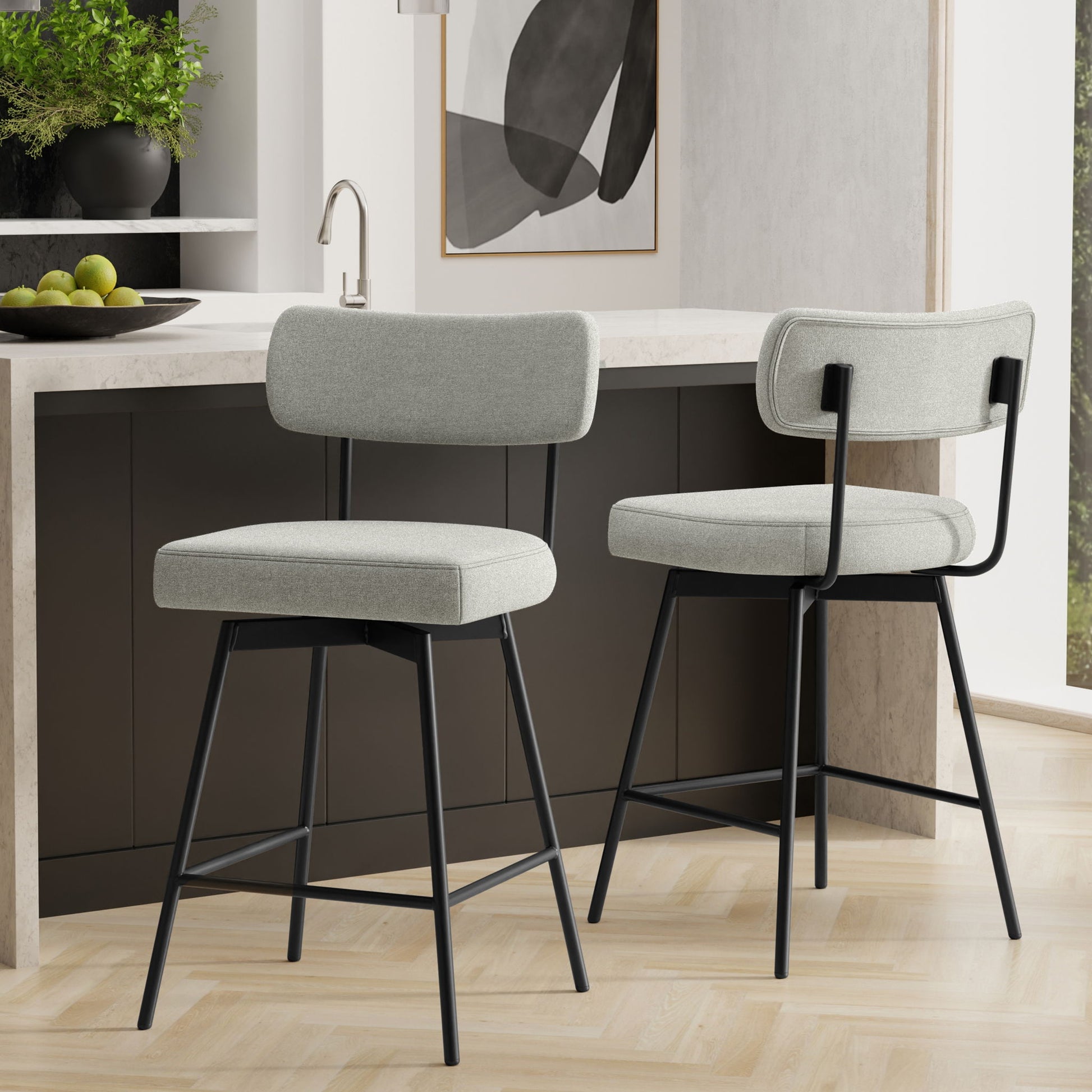 Two Giselle Swivel Counter Height Stools, featuring light gray cushioned seats and backs with black metal legs, are set in a modern kitchen with a dark island, marble countertop, and abstract wall art. Set includes 2 stools.