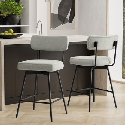 Two Giselle Swivel Counter Height Stools, featuring light gray cushioned seats and backs with black metal legs, are set in a modern kitchen with a dark island, marble countertop, and abstract wall art. Set includes 2 stools.