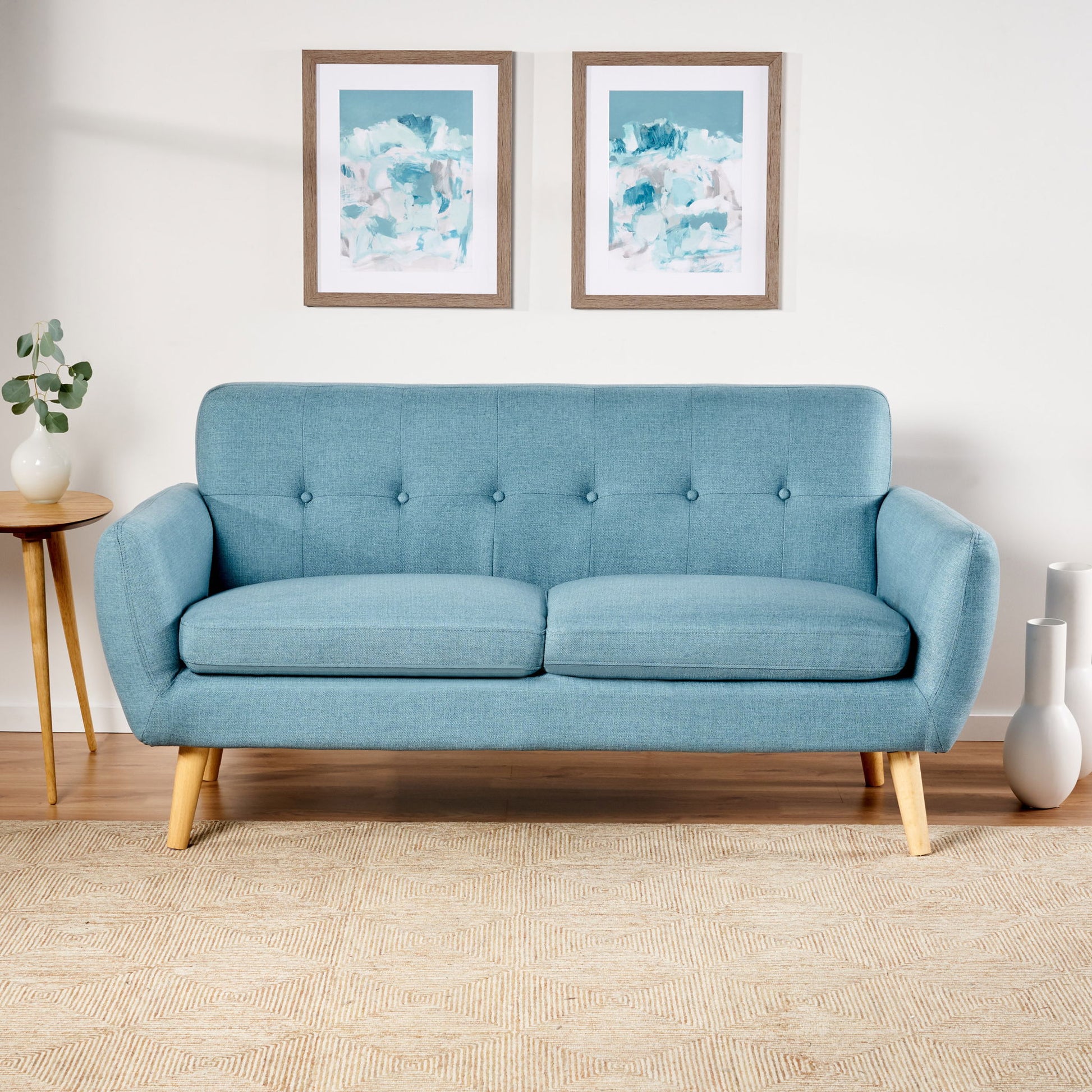 A Modern Loveseat Sofa With Button Tufted Backrest in light blue, featuring wooden legs, sits on a beige rug. To the left is a round wooden side table with a plant, and two abstract blue and white paintings hang above the sofa.