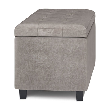The Cosmopolitan Upholstered Storage Ottoman is rectangular, light gray, features subtle top seam detailing, and stands on black wooden legs. Shown from the front angle.