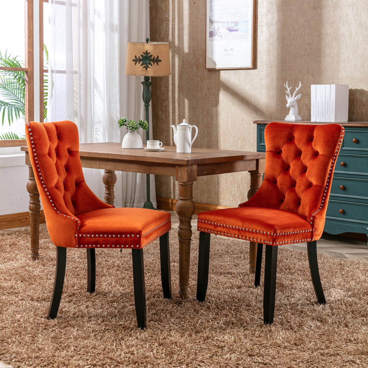 Two Nikki modern tufted velvet dining chairs with wood legs and nailhead trim sit by a wooden table on a beige carpet in a cozy, sunlit dining room with a lamp, blue dresser, and decorative accents in the background.