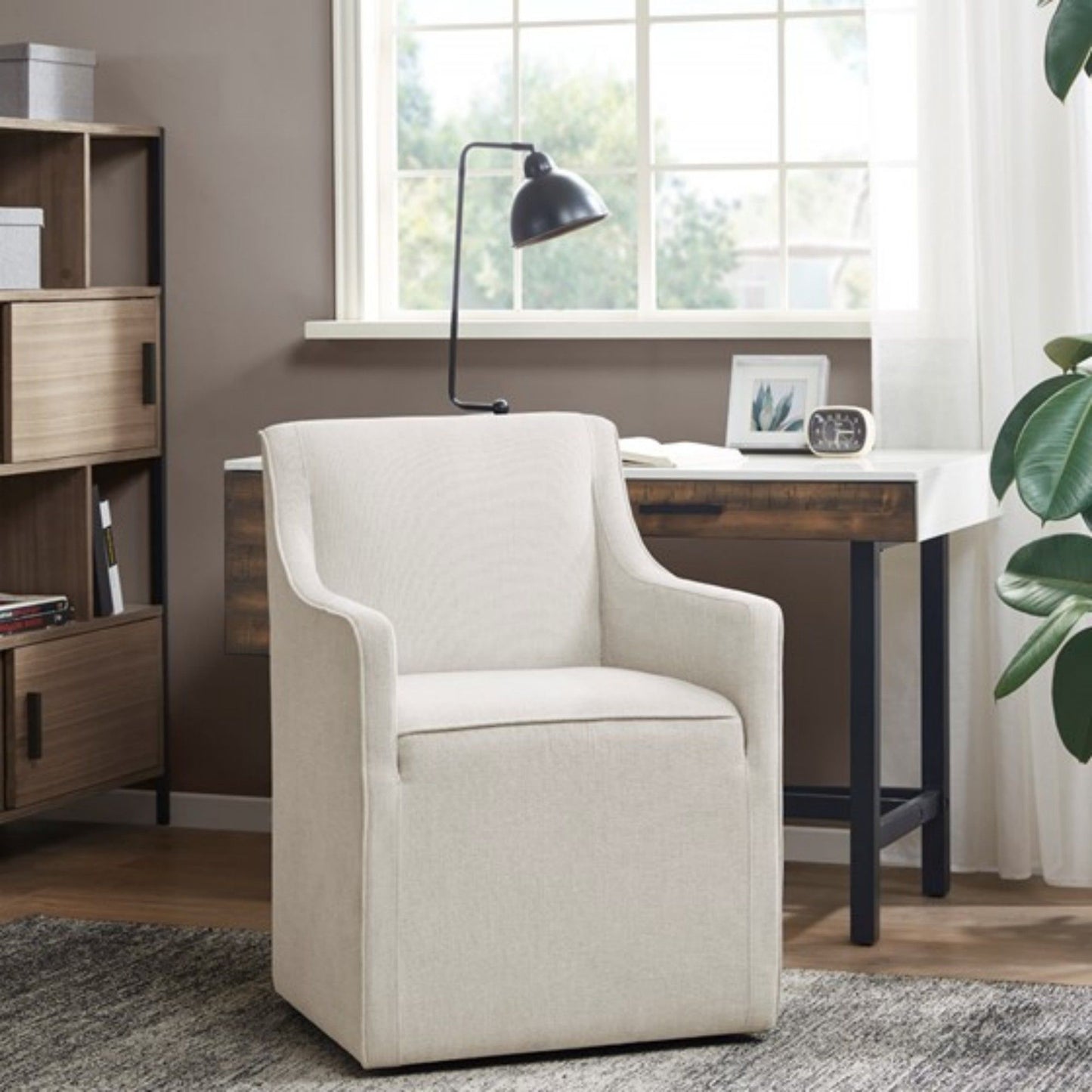 A Traditional Skirted Dining Arm Chair in beige sits by a modern wooden desk near a window, paired with a black lamp, framed art, and bookshelf. Natural light brightens the cozy, contemporary space.