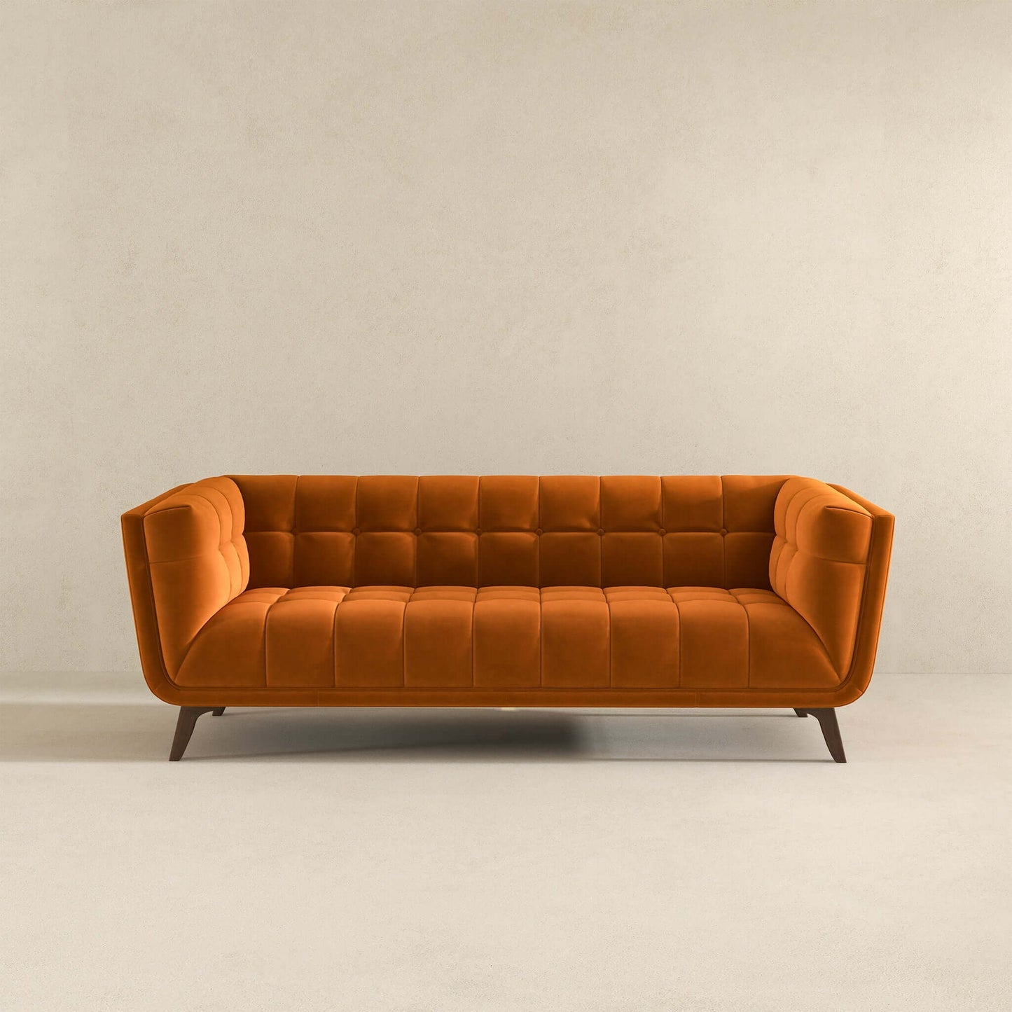 The Addison Mid-Century Modern Design Tufted Sofa in bold orange velvet features a tufted back and seat, angled arms, and wooden legs, displayed against a plain beige wall and floor.