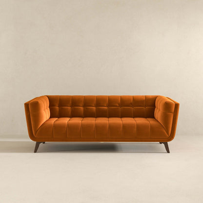 The Addison Mid-Century Modern Design Tufted Sofa in bold orange velvet features a tufted back and seat, angled arms, and wooden legs, displayed against a plain beige wall and floor.