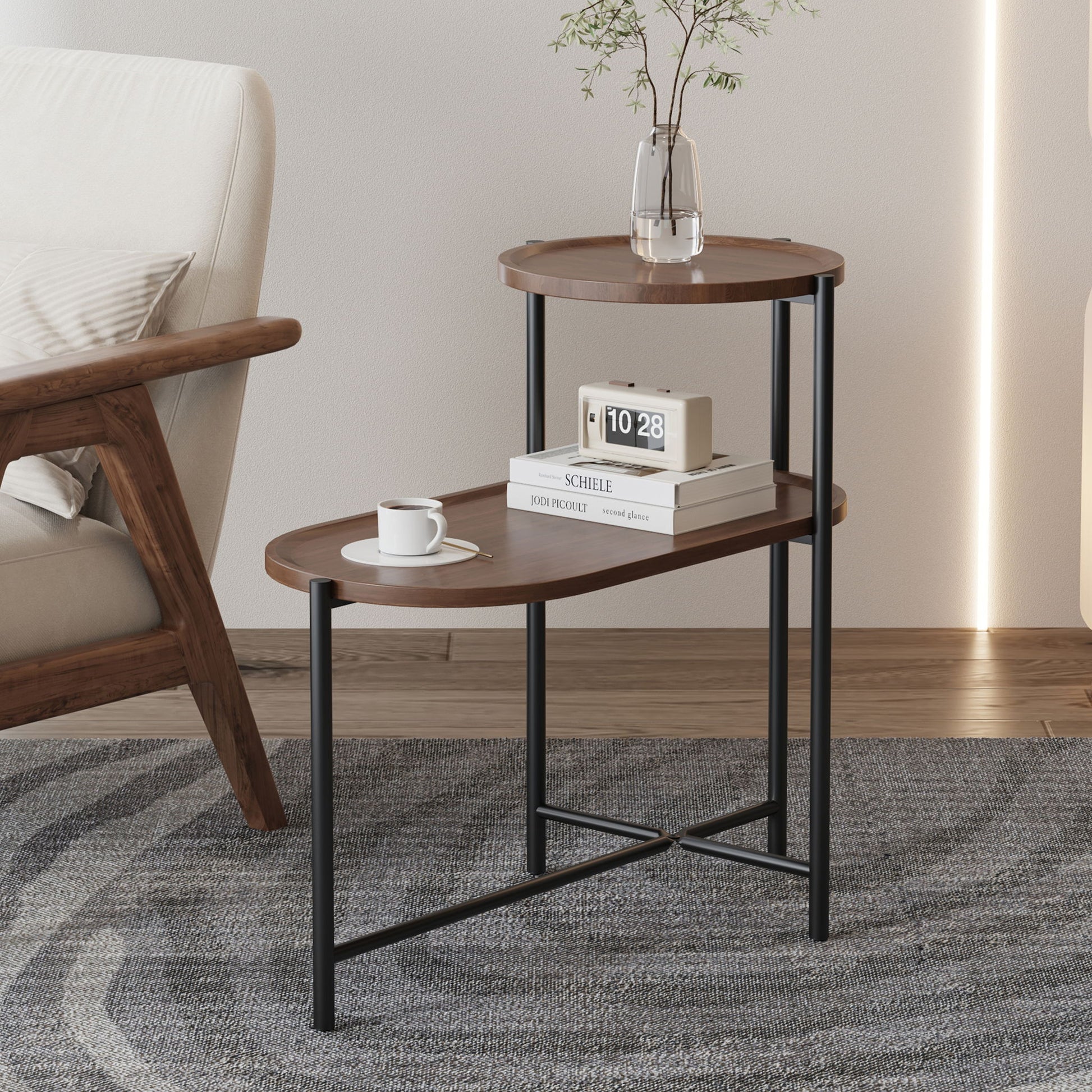 The Round Coffee Table With Storage Tray 2 Tier Oval End Tables Wooden Small Side Table for Living Room, featuring black metal legs, holds a digital clock, book, and vase with flowers beside an upholstered chair on a gray patterned rug.