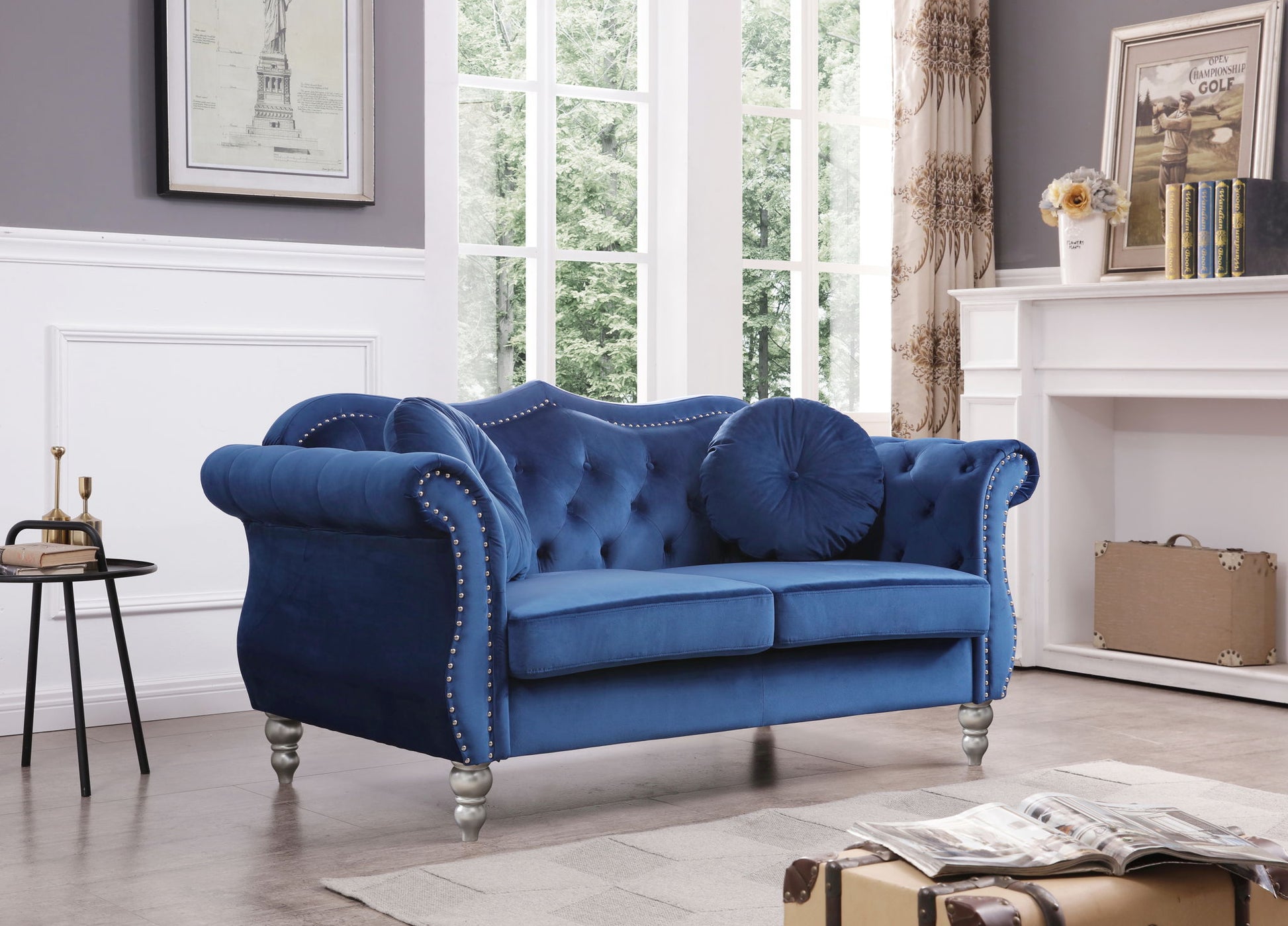 The Stylish Flared Arm Loveseat, featuring blue velvet upholstery, tufted details, silver stud accents, and two round pillows, is showcased in a bright living room with large windows and white paneled walls.