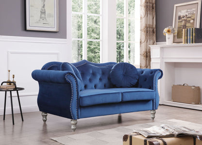 The Stylish Flared Arm Loveseat, featuring blue velvet upholstery, tufted details, silver stud accents, and two round pillows, is showcased in a bright living room with large windows and white paneled walls.
