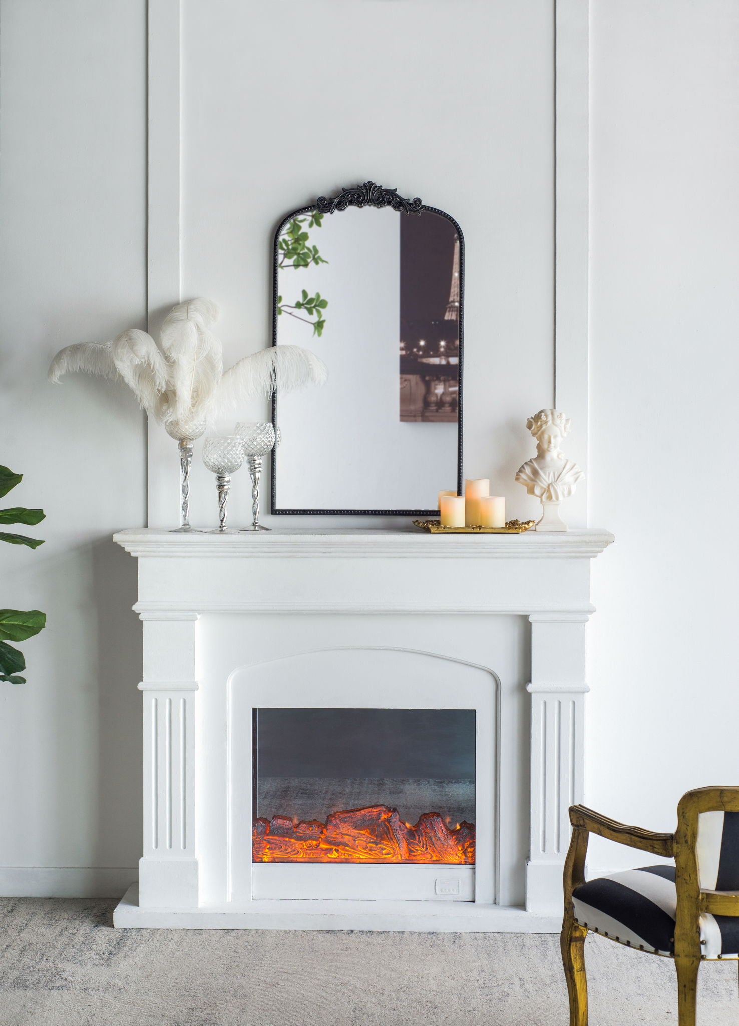 A white fireplace displays a glowing electric fire and is topped by the Arch Mirror, Baroque Inspired Wall Decor. Nearby, candles on a gold tray, white feathers in a vase, a small bust, and a gold-accented chair add elegant touches.