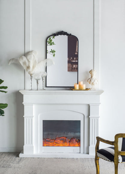 A white fireplace displays a glowing electric fire and is topped by the Arch Mirror, Baroque Inspired Wall Decor. Nearby, candles on a gold tray, white feathers in a vase, a small bust, and a gold-accented chair add elegant touches.