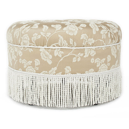 The Yolanda Round Upholstered Accent Ottoman features a beige fabric with white floral patterns, a soft cushioned top, decorative white fringe trim at the base, and offers an elegant vintage look.