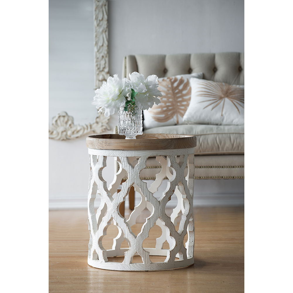 A Stylish Rustic Round Wooden Table holds a vase of white flowers. Behind it sits a beige tufted sofa with decorative pillows and a large mirror with a carved frame on the wall.