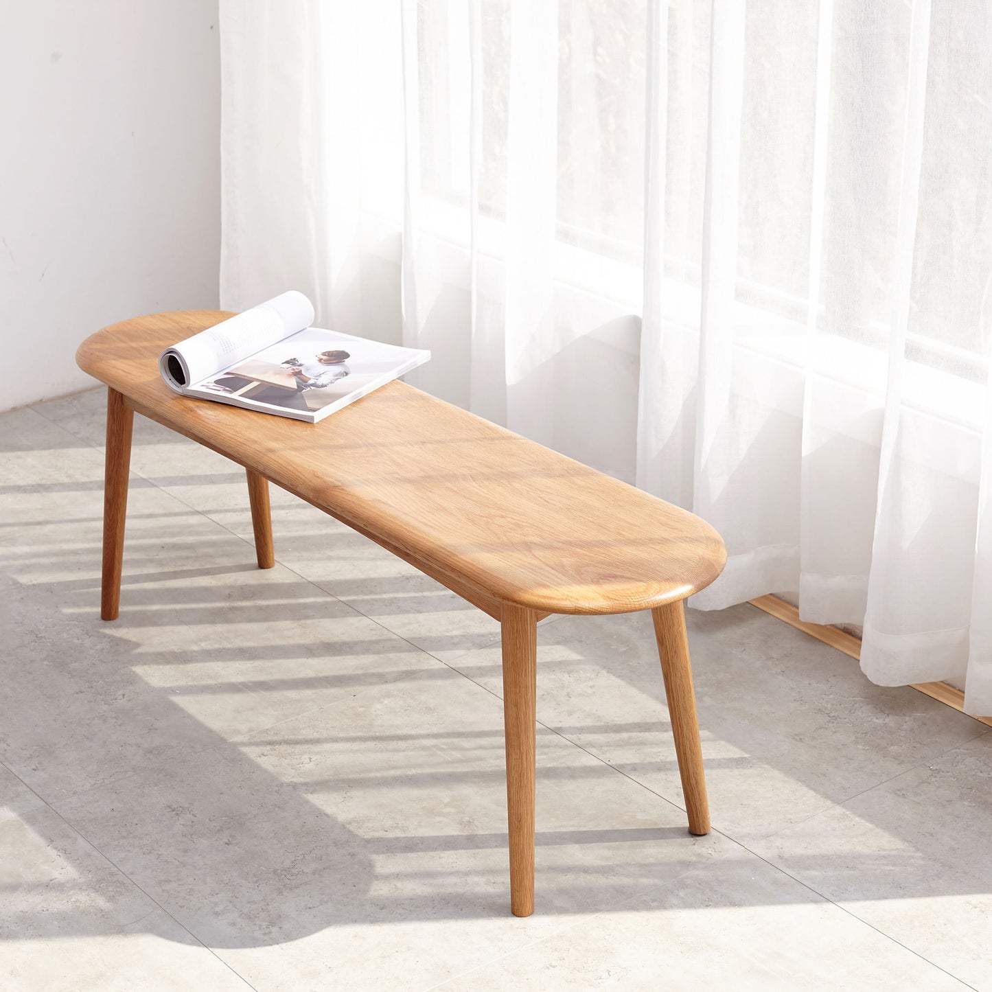 The Dining Bench, Table Bench For Living Room features light wood and rounded edges, positioned on a tiled floor near sheer white curtains. A magazine and rolled-up paper sit atop it, illuminated by natural sunlight.