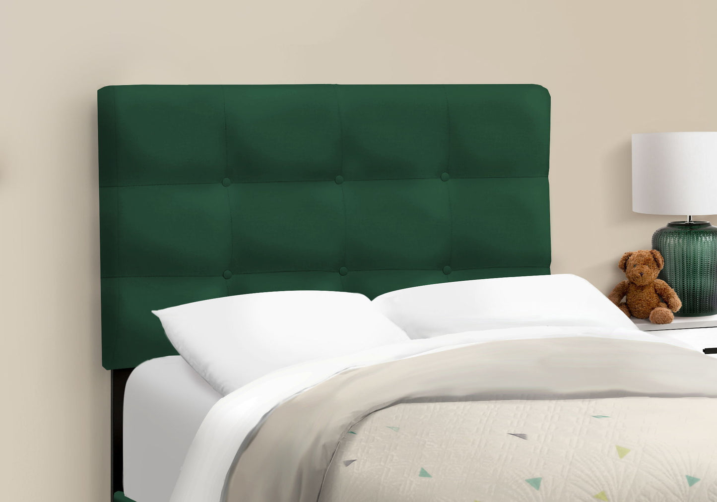 A Bed, Upholstered, Frame Only, Transitional with a dark green tufted headboard is set against a beige wall, featuring white bedding and pillows. A nightstand with a lamp and brown teddy bear stands beside it.