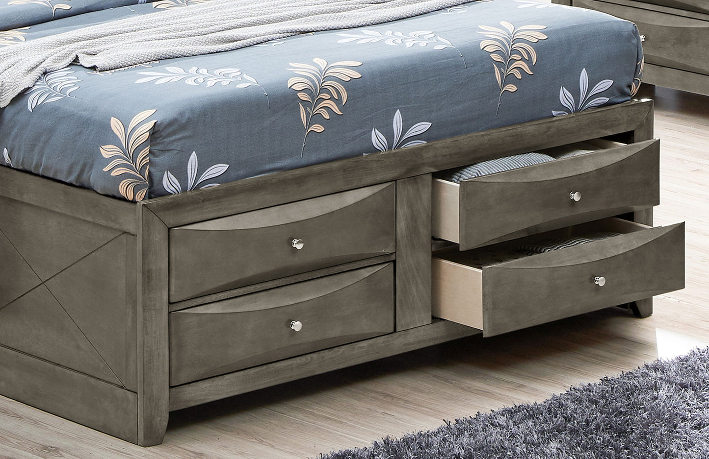 A close-up of the Marilla - Storage Bed With Bookcase Headboard in gray, featuring four built-in drawers—two open to reveal under-bed storage—with a gray and white floral comforter on a light wood floor.