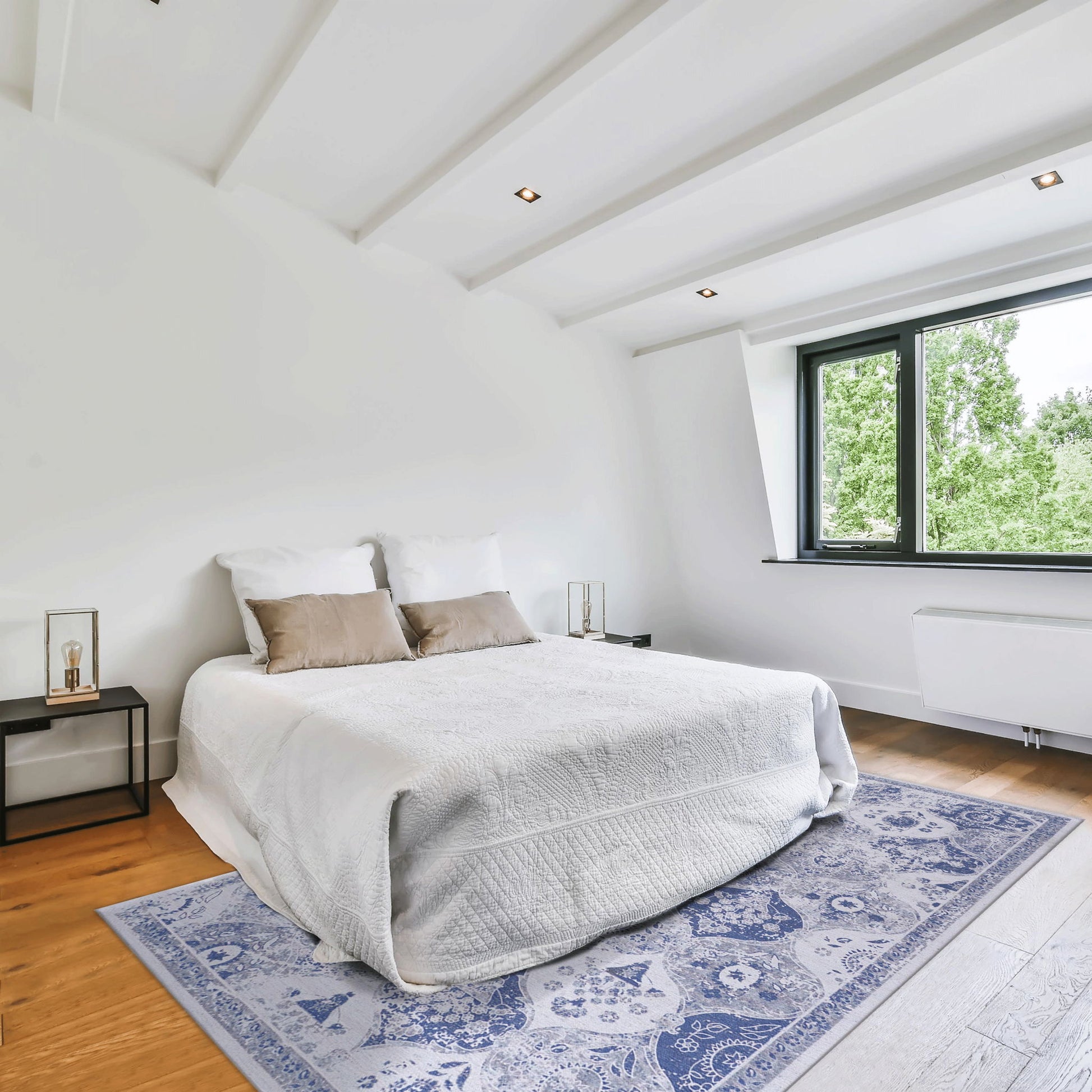 A bright, modern bedroom with a neatly made bed, white bedding, two brown accent pillows, bedside tables with lamps, wooden floor, large window with green trees outside, and a blue Maval - Area Rug adding pattern to the space.