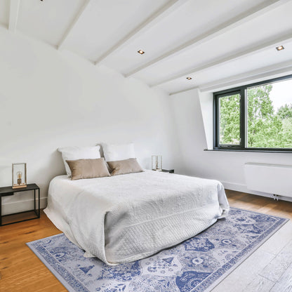 A bright, modern bedroom with a neatly made bed, white bedding, two brown accent pillows, bedside tables with lamps, wooden floor, large window with green trees outside, and a blue Maval - Area Rug adding pattern to the space.