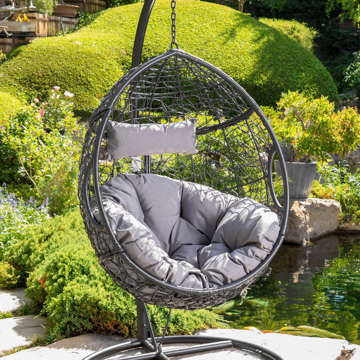 The Kylie Boho Style Hanging Basket Chair, crafted from rattan with water resistant cushions, is placed on a patio by a pond and surrounded by greenery and trimmed bushes in the garden.