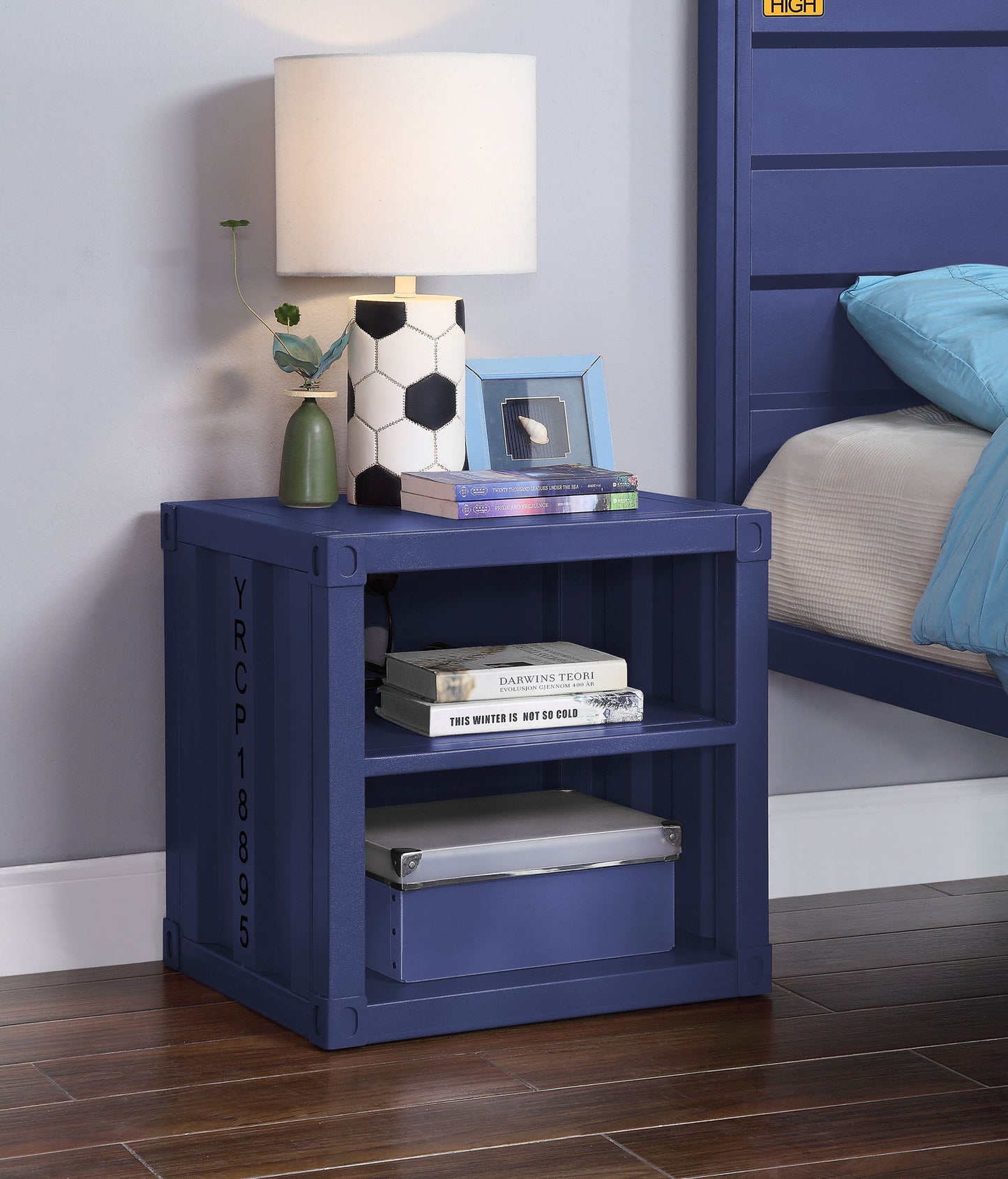 A blue Cargo Nightstand With USB sits on a wooden floor beside a bed, holding a lamp, hexagonal vase with plant, two books, picture frame, and storage box. It features an industrial design for modern bedrooms.