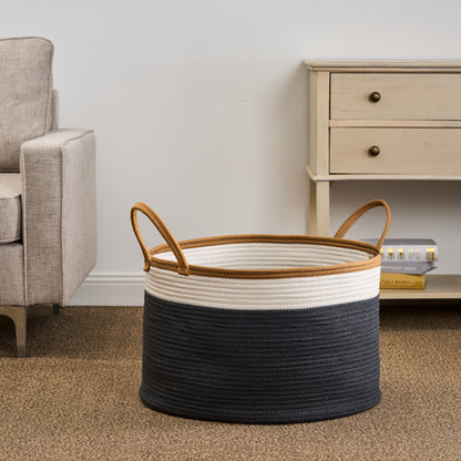 The Francis - Cotton Round Laundry And Storage Basket, featuring brown handles and a black and white color-block design, sits on a beige carpet beside a light armchair and cream-colored side table.