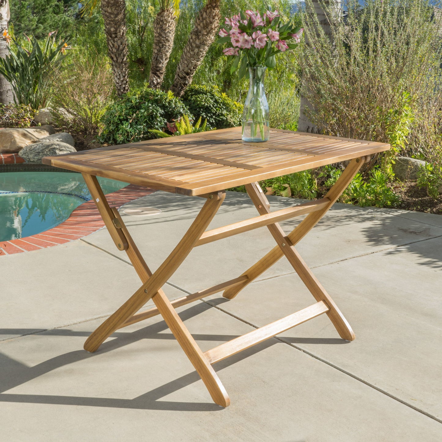 The Blissique Outdoor Foldable Wood Dining Table with a slat design and crossed legs stands on concrete by a pool, topped with a glass vase of pink flowers. Lush greenery and palm trees fill the background.