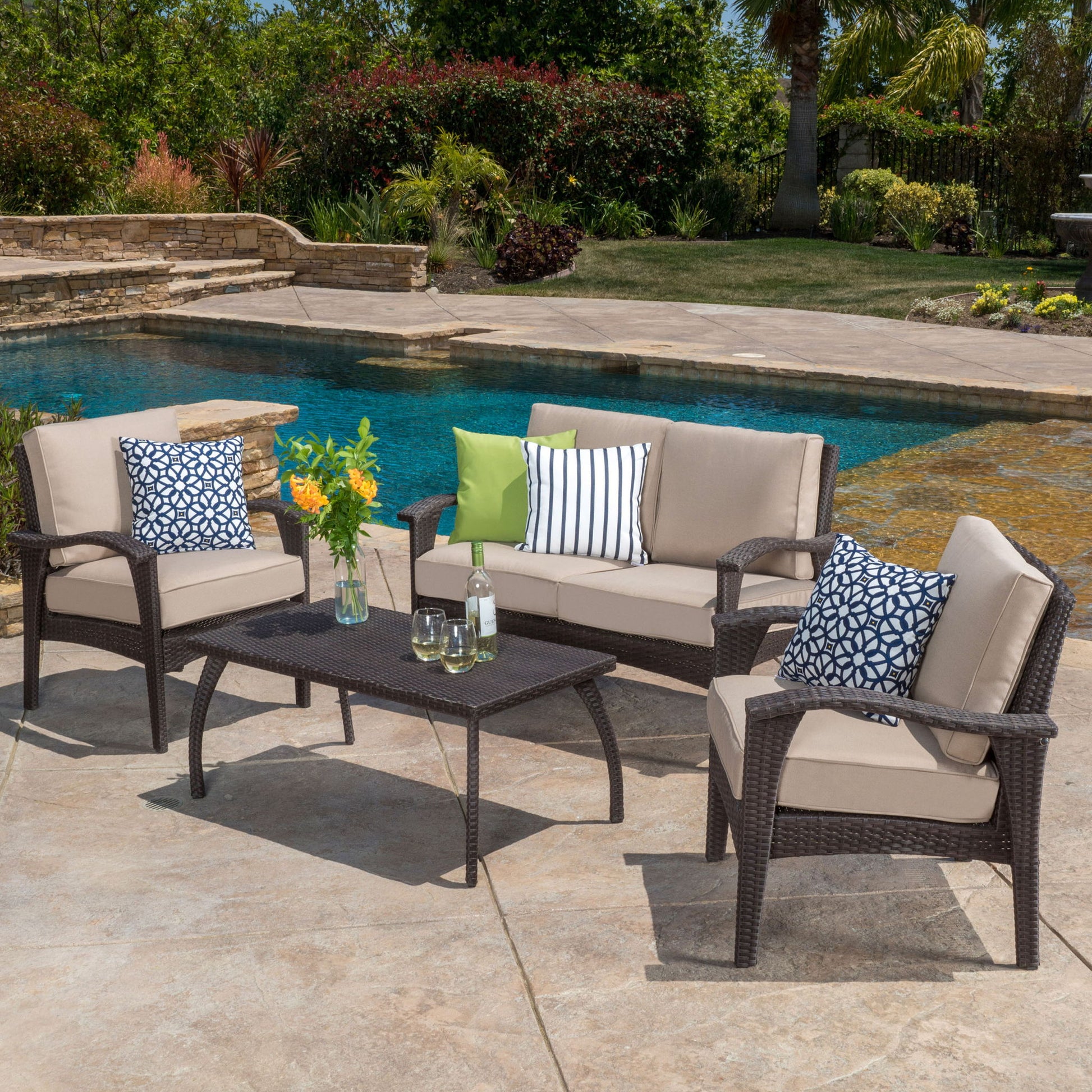Outdoor patio with pool featuring the Honolulu - Wicker Set Outdoor Seating: a wicker sofa, two chairs with beige cushions, a coffee table with glassware, and decorative pillows in black, white, green, and blue patterns.