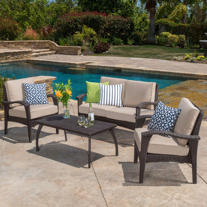 Outdoor patio with pool featuring the Honolulu - Wicker Set Outdoor Seating: a wicker sofa, two chairs with beige cushions, a coffee table with glassware, and decorative pillows in black, white, green, and blue patterns.