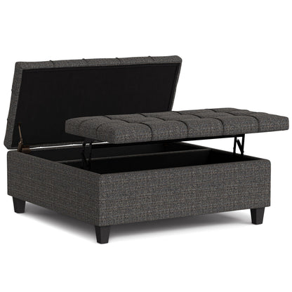 The Harrison Upholstered Coffee Table Storage Ottoman features a gray tufted, hinged lid, open to show its empty storage compartment. It stands on four short black legs.